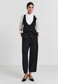 Donna in piedi con un gilet nero abbottonato con spilla floreale, camicia bianca trasparente, pantaloni neri a culotte e tacchi slingback neri.