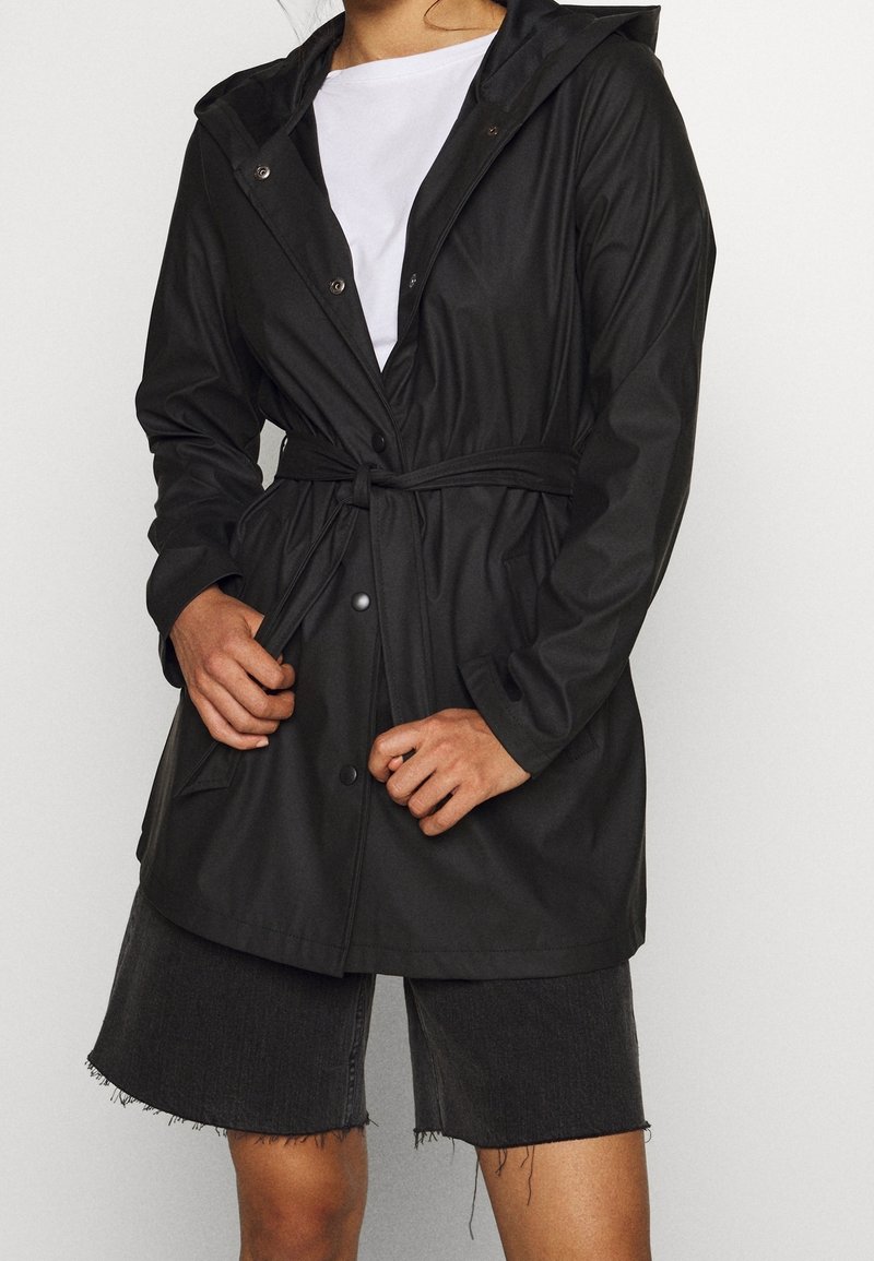 Veste noire imperméable avec capuche, ceinture élastique à la taille, boutons-pression et poches latérales, associée à un short en jean foncé.