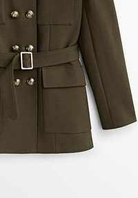 Veste vert olive avec un devant à double boutonnage, des accents de boutons dorés, une taille ceinturée et deux poches avant. Texture de tissu lisse.