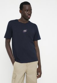 Camiseta de algodón azul marino con mangas cortas, con un logo rectangular en blanco y morado en el área del pecho izquierdo.