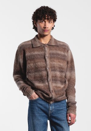 Braun gestreifter Cardigan aus einem flauschigen Material, mit Kragen, sechs Knöpfen und gerippten Bündchen, kombiniert mit blauen Jeans.