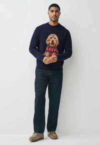 Granatowy sweter z teksturowanym, wielokolorowym wzorem psa w deseń argyle na piersi, w zestawieniu z ciemnymi dżinsami i beżowymi butami.