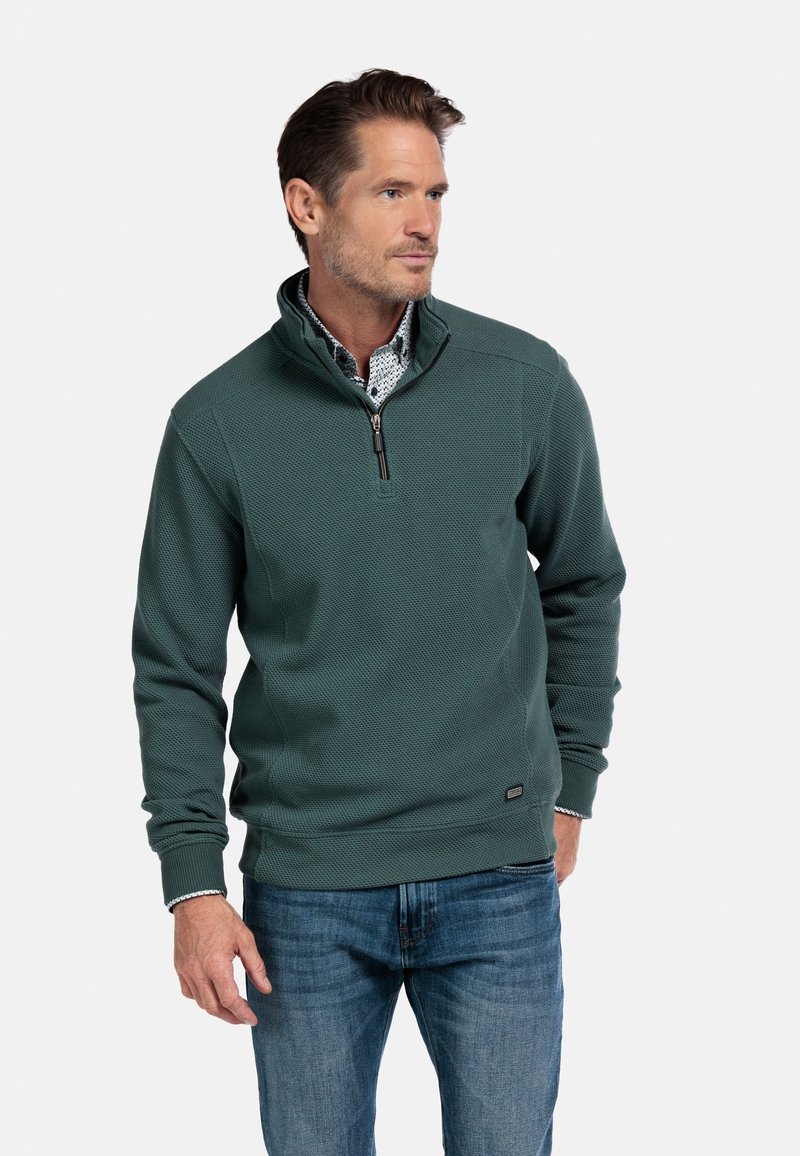 Groene gestructureerde trui met een ritskraag, voorzien van lange mouwen en een subtiel patroon. Gepaard met een blauwe spijkerbroek, staande tegen een effen achtergrond.