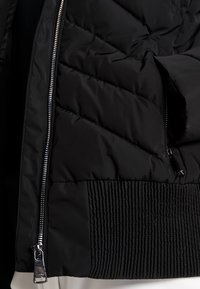 Gros plan d'une veste noire matelassée avec des coutures diagonales, une fermeture éclair argentée et une ceinture côtelée en tissu cousu.