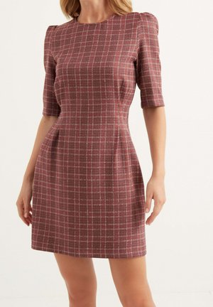 Kobieta w dopasowanej, bordowej, kraciastej sukience do kolan z rękawami do łokci na jednolitym tle.