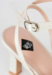 Sandale blanche à talons hauts avec une sangle texturée, boucle en argent et semelle intérieure rose poudré. Talon carré et étiquette de la marque visibles.
