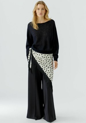 Frau trägt einen schwarzen flauschigen Pullover, weit geschnittene schwarze Hose und ein weißes Tupftuch, das um die Taille gebunden ist.