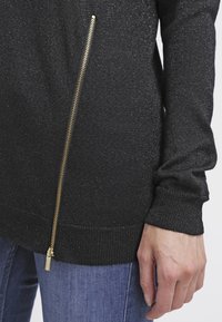 Schwarzer Pullover mit metallic-finish, seitlichem Reißverschlussdetail in Gold und geripptem Saum. Weicher Stoff, enganliegende Ärmel.