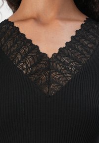 Abito nero a coste con scollo a V e un delicato strato di pizzo, che mette in mostra intricati motivi lungo il collo.