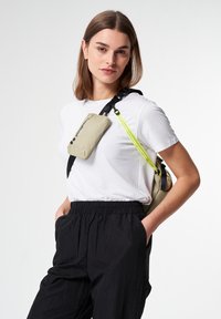 Frau mit glattem braunem Haar, die ein weißes T-Shirt und schwarze Hosen trägt, während sie eine beige Umhängetasche mit gelbem Schulterriemen über der Schulter trägt.