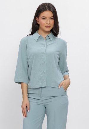 Kvinde med langt mørkt hår iført en lyseblå skjorte med knapper og trekvartærmer samt matchende bukser, stående med den ene hånd i lommen.