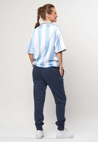 Žena s ispletenim repom okrenuta je leđima, nosi plavo-bijelu prugastu majicu, tamnoplave hlače za trčanje, bijele tenisice, zlatne naušnice u obliku obruča i narukvicu.