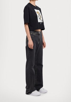 Personne portant un t-shirt court noir avec un graphisme d'ours, un jean noir ample et des baskets blanches, debout devant un fond blanc.