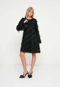 Μαύρο φόρεμα με υφή από υλικό που διαθέτει στρώματα φουντωτής επιφάνειας, φαρδιά εφαρμογή, μακριά μανίκια και μήκος μέχρι το γόνατο; συνδυασμένο με γυαλιστερές μαύρες γόβες.