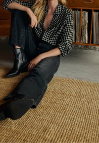 Personne assise par terre portant une chemise à carreaux noire, un jean noir et des bottes en cuir noires près d'une étagère en bois avec des disques vinyles.