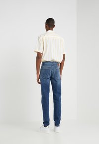 Keltainen ja valkoinen raitapaita lyhyillä hihoilla yhdistettynä sinisiin farkkuihin, jotka ovat standardimallisia ja joissa on takataskut.