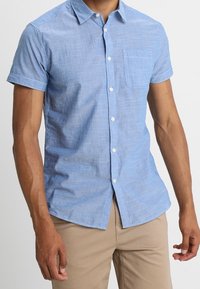 Mężczyzna ubrany w jasnoniebieską koszulę na krótki rękaw z kieszenią na piersi oraz beżowe spodnie, stojący na neutralnym tle.