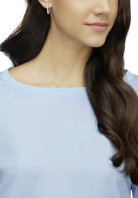 Srebrne kolczyki koła z fakturowaną powierzchnią, noszone przez osobę w jasnoniebieskiej bluzce z okrągłym dekoltem. Widać długie, falowane ciemne włosy.