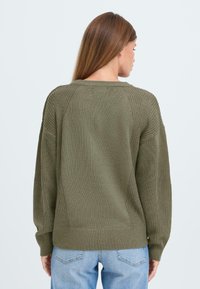 Olivgrön tröja med texturerad stickning, ribbade ärmslut och nederkant, med en lös passform och sänkta ax seams, tillsammans med ljusblå jeans.