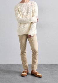 Kremowy sweter w warkocze z prążkowanymi ściągaczami, jasnobeżowe chinosy oraz brązowe zamszowe mokasyny z kremowym akcentem. Widoczne detale tekstury materiału.