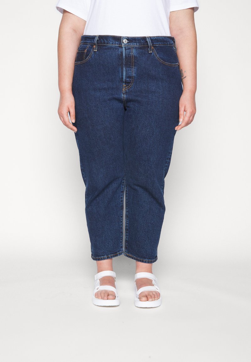 Dunkelblaue Denim-Hose im geraden Schnitt, mit fünf Taschen, Knopfverschluss und kürzerer Länge. Tragen mit weißen Sandalen.