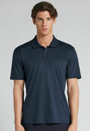 Junger Mann mit hellem Teint und welligem braunem Haar, der ein dunkelmarineblaues Polohemd mit kurzen Ärmeln vor einem schlichten Hintergrund trägt.