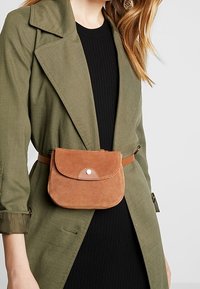 Bruine suède heuptas met een afgeronde klep en metalen drukknopdetail, gedragen over een olijfgroene jas met een slanke bruine riem.