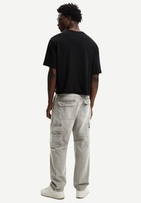 Muškarac nosi crnu majicu, svijetlosive cargo hlače s džepovima, bijele tenisice, stoji okrenut leđima na jednostavnoj bijeloj pozadini.