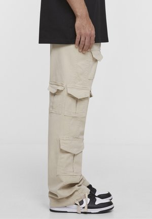 Oldalnézet egy személyről, aki bézs rakott nadrágot visel több zsebfedővel, fekete-fehér tornacipővel és fekete pólóval.