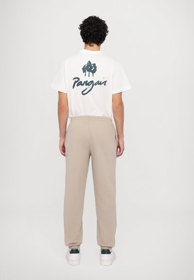 Hvid bomulds t-shirt med grønt logo på ryggen; beige sweatpants med elastiske manchetter; modellen har hvide sneakers på.