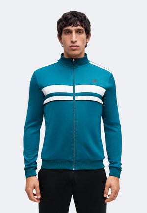 Hombre con chaqueta de cremallera color azul verdoso con rayas horizontales blancas en el pecho y pantalones negros, posando sobre un fondo liso.