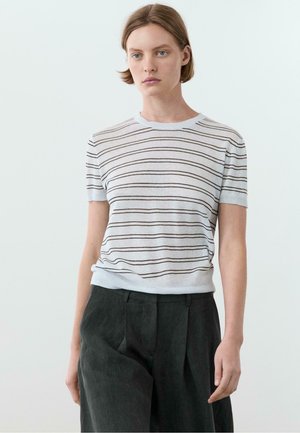 Ung person iført en hvid kortærmet skjorte med tynde vandrette striber og mørke, løstsiddende bukser, stående mod en ensfarvet baggrund.