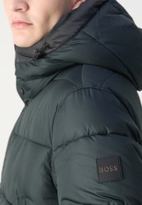 Mørkegrønn polstret jakke med høy krage, med hette i svart og synlig sømdesign; inkluderer et lærmerket BOSS.