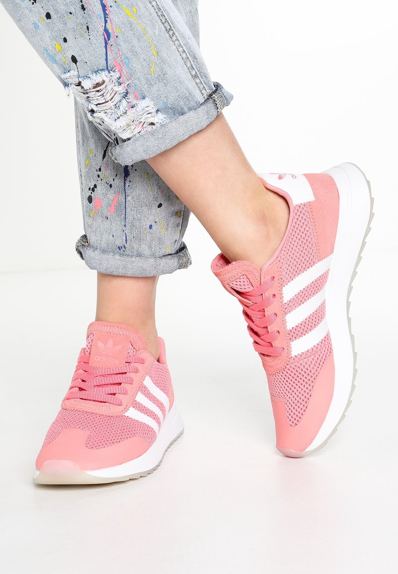 Rosa träningsskor med vita ränder och nätdesign. Sålen är vit och ovandelen har en texturerad yta. Rivna jeans med färgsplatter.