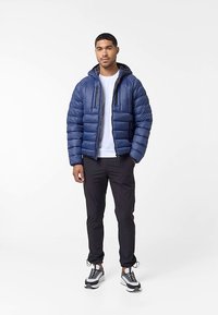 Άντρας που στέκεται φορώντας ένα μπλε μπουφάν, λευκό μπλουζάκι, μαύρα παντελόνια και μαύρα-άσπρα αθλητικά παπούτσια μπροστά σε απλό λευκό φόντο.