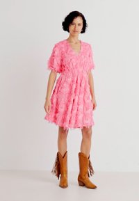 Ροζ φόρεμα με λουλουδάτη υφή και ρέουσες λεπτομέρειες, V λαιμόκοψη, κοντά μανίκια και φουσκωτό στρίφωμα, συνδυασμένο με καφέ καουμπόικες μπότες με φούντες.