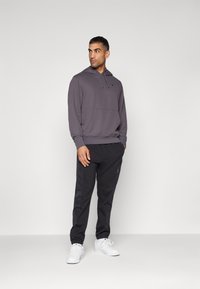 Felpa grigia con cappuccio e tasca frontale, abbinata a pantaloni sportivi neri a sigaretta con tasche laterali e scarpe da ginnastica bianche.
