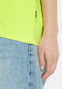 Neon-gelbes Baumwoll-T-Shirt mit einem Seitenschild "SENSES", kombiniert mit blauen Denim-Jeans, die eine Steppnaht an der Tasche zeigen.