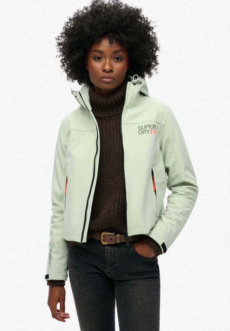 Veste vert menthe avec un design ajusté, capuche, poches zippées et accents noirs. Portée par-dessus un pull en tricot épais brun.