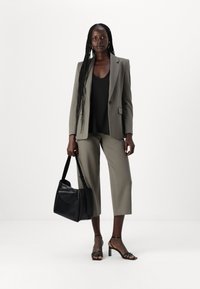 Grå to-delt dress med korte bukser, en svart topp og svarte sandaler med stropper. Holder en svart håndveske. Ensfarget hvit bakgrunn.