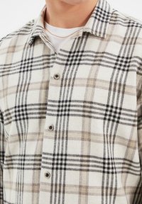 Camicia a quadri nei colori beige, nero e bianco. Tessuto morbido con una trama in flanella. Presenta un colletto, una chiusura frontale con bottoni e motivi a quadri a contrasto.