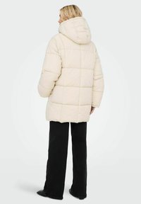 Chaqueta de plumas crema con capucha, que presenta un diseño acolchado. Combinada con pantalones de pierna ancha negros, el conjunto se encuentra frente a un fondo claro.