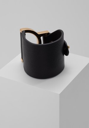 Bracelet large en cuir noir avec fermoir en métal doré, présenté sur un cube blanc sur fond gris.