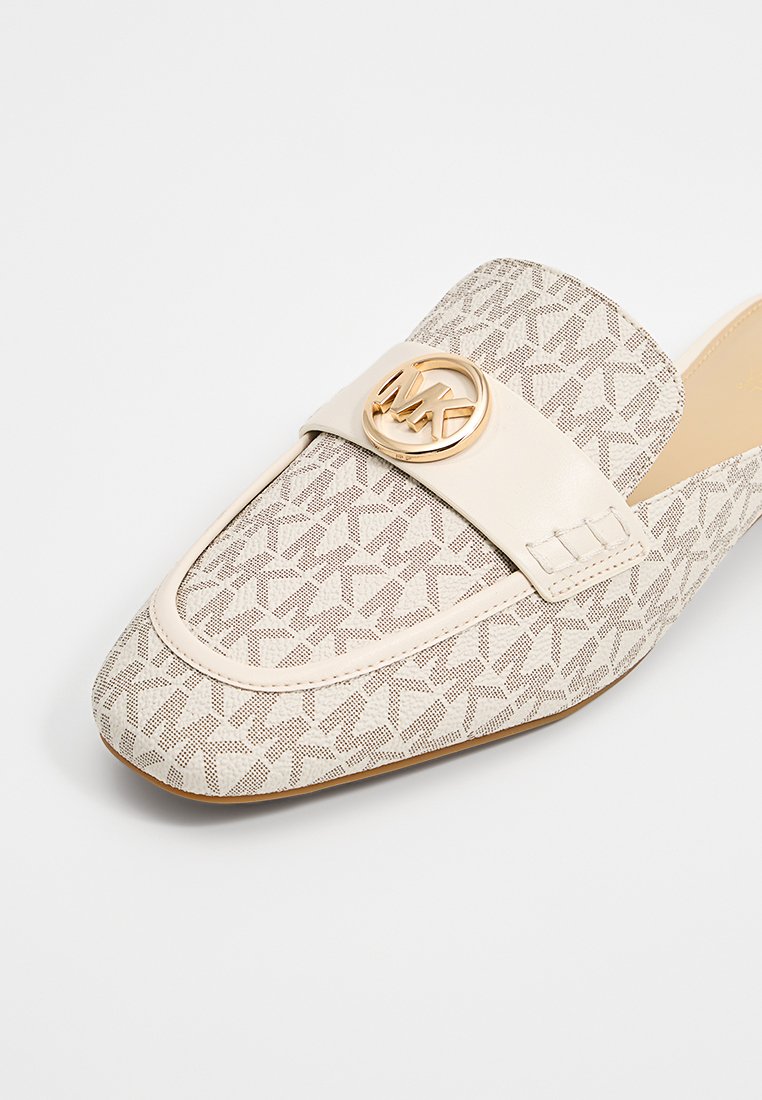 Beige instap schoen met een geometrisch patroon, voorzien van een gouden cirkelvormig logo en een gestructureerde leren band over de bovenkant. Plat zool.