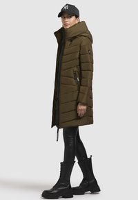 Manteau rembourré vert olive avec un col haut, fermeture éclair, et poches zippées sur les côtés, associé à des leggings noirs et des bottes montantes noires à lacets.