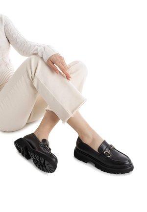 Mocasines de cuero negro con una suela gruesa y herrajes decorativos en dorado. Los zapatos presentan un acabado liso y una suela con un dibujo detallado.