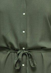 Φόρεμα χακί με κουμπιά και υφασμάτινη ζώνη δεμένη σε φιόγκο στη μέση, που δείχνει λεπτομέρειες και ραφές στην κοιλιακή περιοχή.