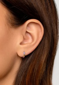 Piccole orecchini a cerchio con pietre preziose multicolori, montati su un metallo argentato, esposti su una pelle liscia e capelli castani.