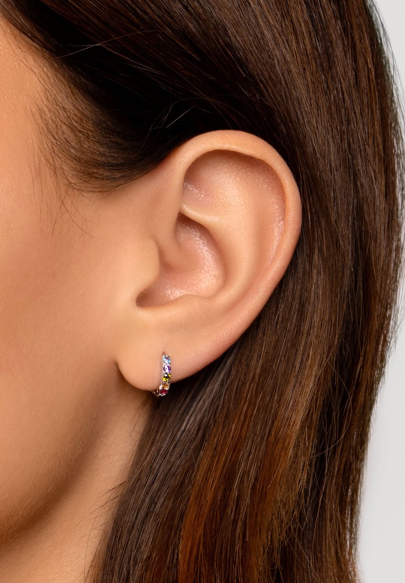 Piccole orecchini a cerchio con pietre preziose multicolori, montati su un metallo argentato, esposti su una pelle liscia e capelli castani.