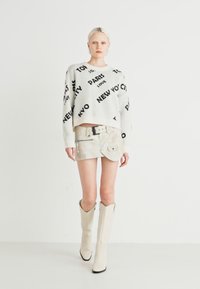 Lys grå strikkegenser med svarte bynavn, sammen med et kort beige skjørt i falsk lær med klaffelomme og beltedetalj. Hvite knehøye støvler.